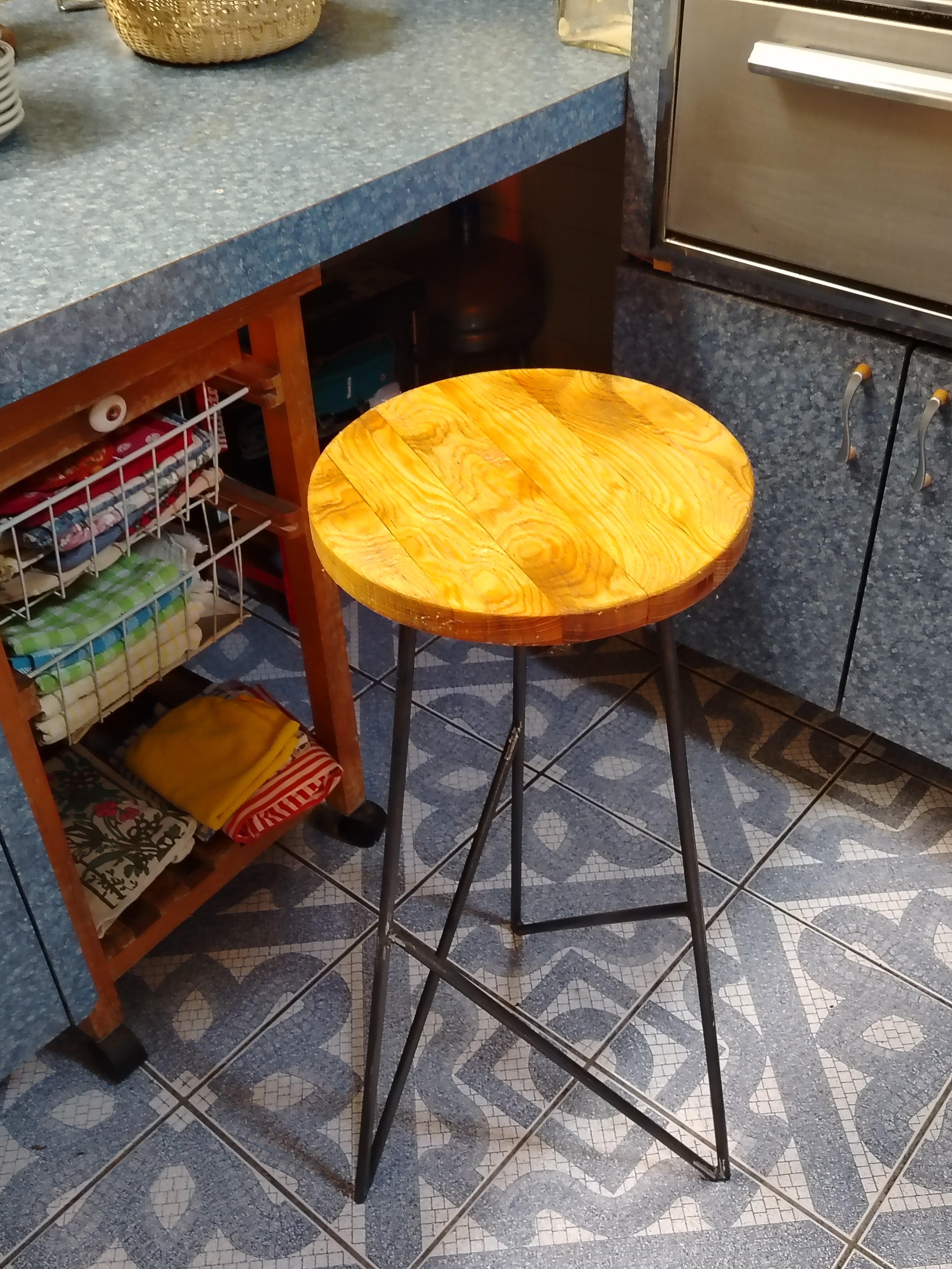 Banco de bar con asiento circular de madera natural y patas delgadas de acero tipo hairpin, en cocina con piso de mosaico