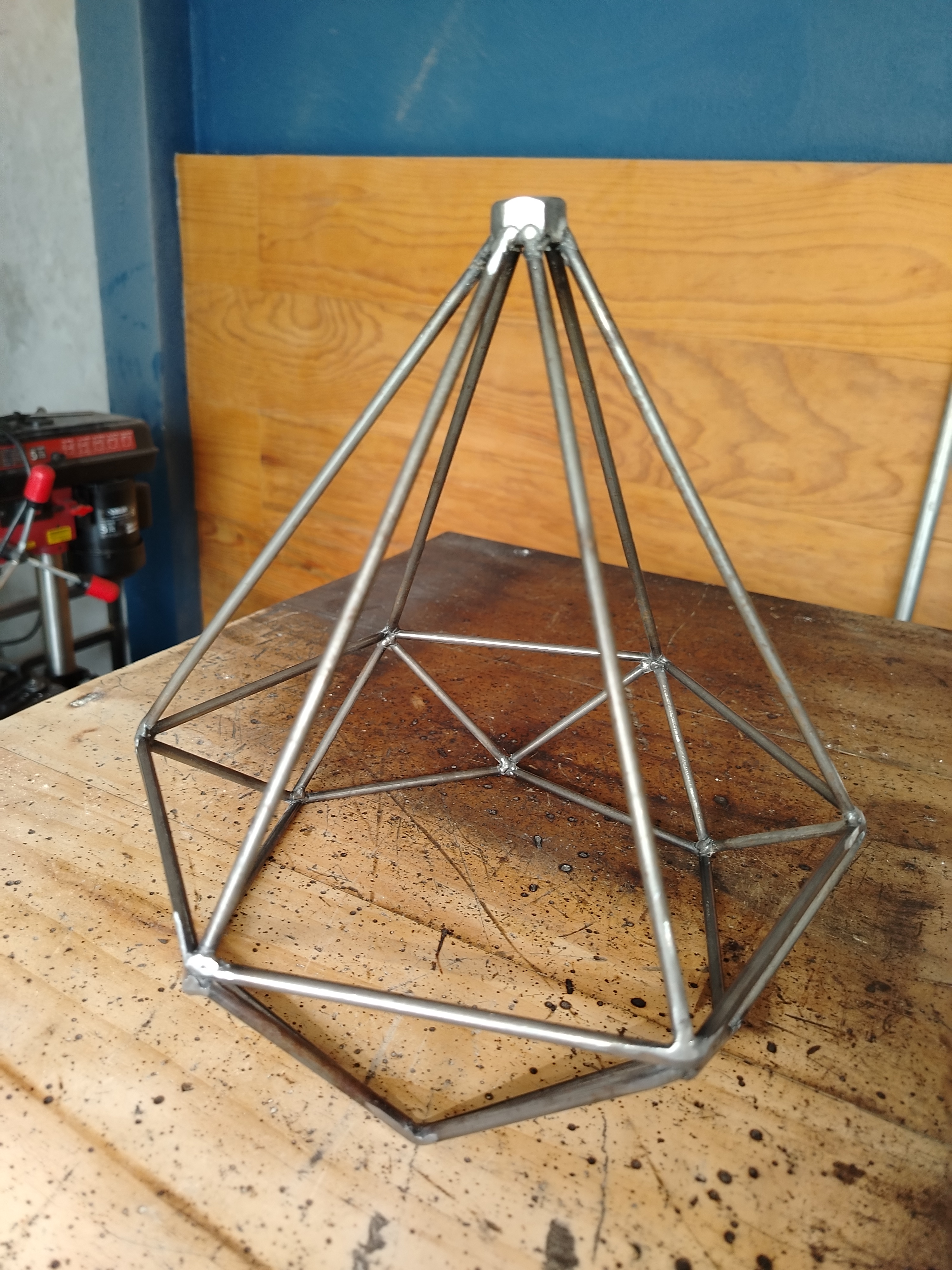 Estructura de lámpara geométrica tipo diamante soldada en varilla de acero inoxidable, sobre mesa de trabajo en taller