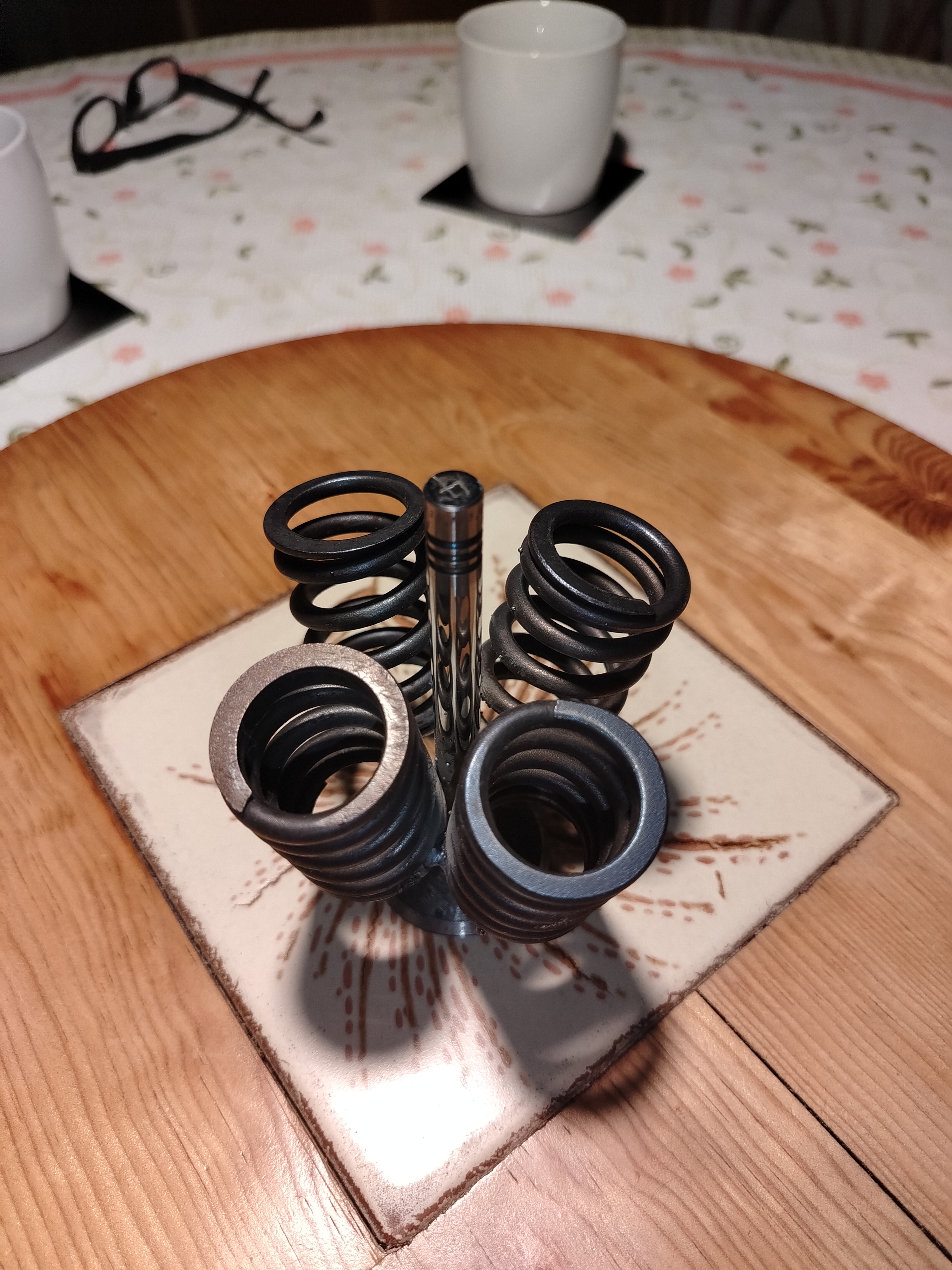 Objeto funcional escultórico hecho con resortes de válvulas de motor y eje central, sobre base de cerámica en mesa de madera